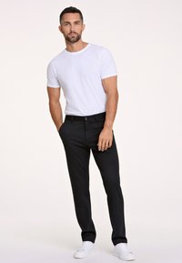 Schwarze Hosen mit schmaler Passform, kombiniert mit einem eng anliegenden weißen T-Shirt mit Rundhalsausschnitt. Das Modell steht selbstbewusst in weißen Turnschuhen.