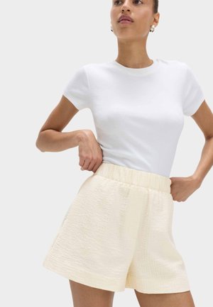 Kvinde iført en tætsiddende hvid kortærmet top og teksturerede cremefarvede shorts med høj talje, stående med hænderne nær taljen.