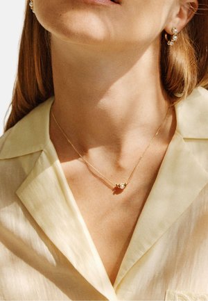 Mujer con blusa crema, collar dorado y pendientes dorados a juego, ambos con detalles de perlas, mostrada desde el cuello hasta los labios.