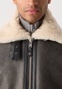Τζάκετ με γιακά από shearling σε γκρι υφασμάτ�ινο υλικό, με μαύρο φερμουάρ, ασημένιες λεπτομέρειες από μέταλλο και μαύρες δερμάτινες λεπτομέρειες.