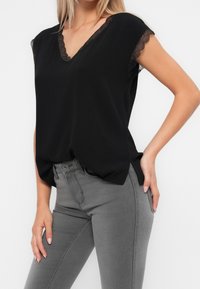 Zwarte V-hals top met kantafwerking bij de mouwen, zachte stof, relaxte pasvorm, gecombineerd met strakke grijze jeans. Zijsplitten aan de onderkant voor bewegingsgemak.