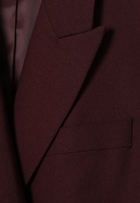 Burgundský blazer s klopovým límcem, texturovanou tkaninou a jednou náprsní kapsou, s hladkou podšívkou viditelnou na pozadí.