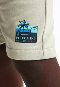 Lichtbeige katoenen shorts beschikken over een zwart en blauw logo-embleem met een palmtree, zon en golven. Afgewerkt met een bijgesneden zoom.
