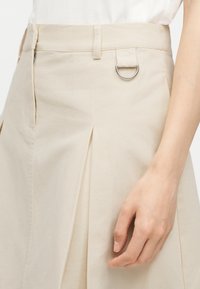 Jupe beige taille haute avec plis sur le devant, passants de ceinture, détail anneau métallique en D, et une main partiellement visible posée à côté.