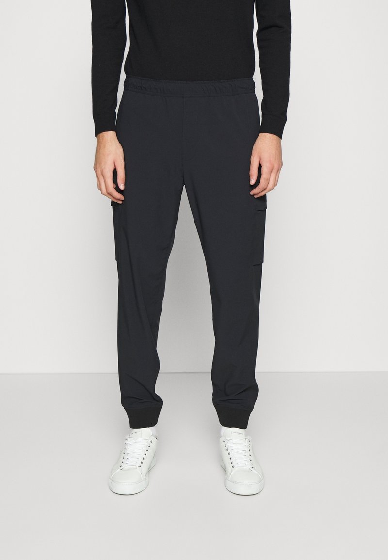 Pantalon cargo noir à taille élastique avec deux poches latérales, poignets ajustés et tissu doux et lisse. Associé à des baskets blanches.