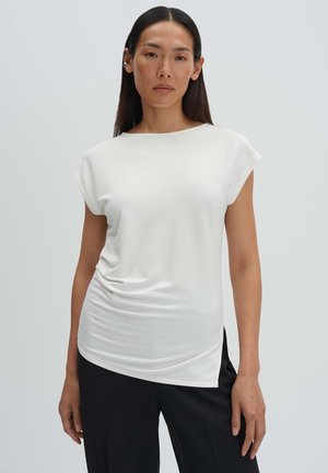 Vrouw met lang donker haar, gekleed in een wit topje met korte mouwen en zwarte broek, staand tegen een eenvoudige lichtgrijze achtergrond.