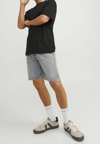 Maglietta nera a maniche corte, pantaloncini di jeans azzurri, calzini bianchi e sneakers beige con dettagli neri. Modello in piedi con una mano sul mento.