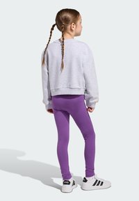 Szara skrócona bluza, fioletowe legginsy i białe tenisówki z czarnymi paskami. Włosy w warkocze, luźny krój na neutralnym tle.