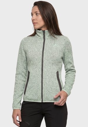 Frau trägt eine hellgrüne Fleecejacke mit Reißverschluss und zwei seitlichen Reißverschlusstaschen, schaut nach links und steht vor einem einfarbigen Hintergrund.