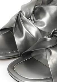 Sandali metallici grigi con cinturini sintetici lucidi intrecciati, design con punta aperta e suola piatta con dettagli di cucitura visibili.