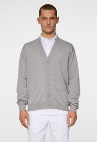 Ljust grå cardigan med v-ringad hals, fem knappar fram och ribbade muddar. Bärs över en vit polo skjorta mot en enfärgad bakgrund.