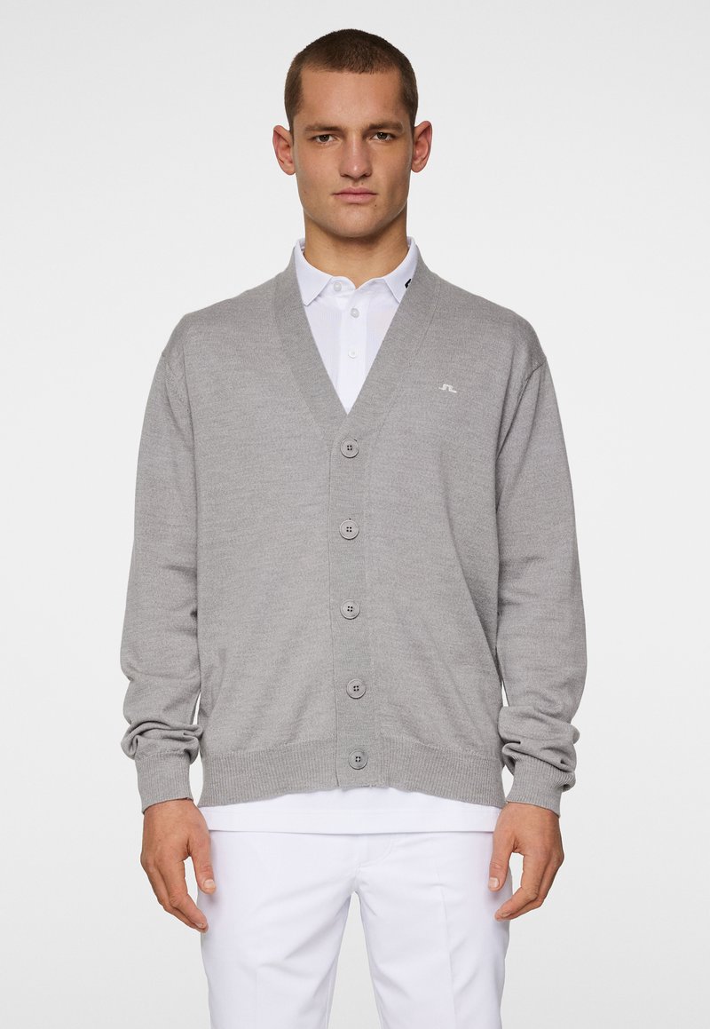 Ljust grå cardigan med v-ringad hals, fem knappar fram och ribbade muddar. Bärs över en vit polo skjorta mot en enfärgad bakgrund.