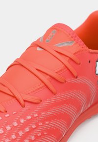 Lyserød koralfarvet træningssko med tekstureret overdel, flade snørebånd og sølvfarvede detaljer. Har et strømlinet design for forbedret ydeevne.