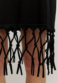 Schwarzes Kleid mit einem verknoteten Fransen-Detail am Saum, das von oben bis unten in einem Diamantmuster gebunden ist, aus weichem Stoff gefertigt.