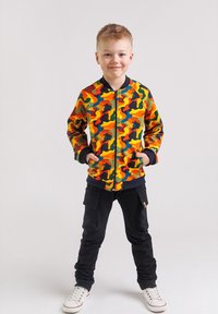 Chłopiec stoi z rękami w kieszeniach, ubrany w kolorową kurtkę w kamuflaż, czarne spodnie cargo i białe sneakersy na jednolitym tle.