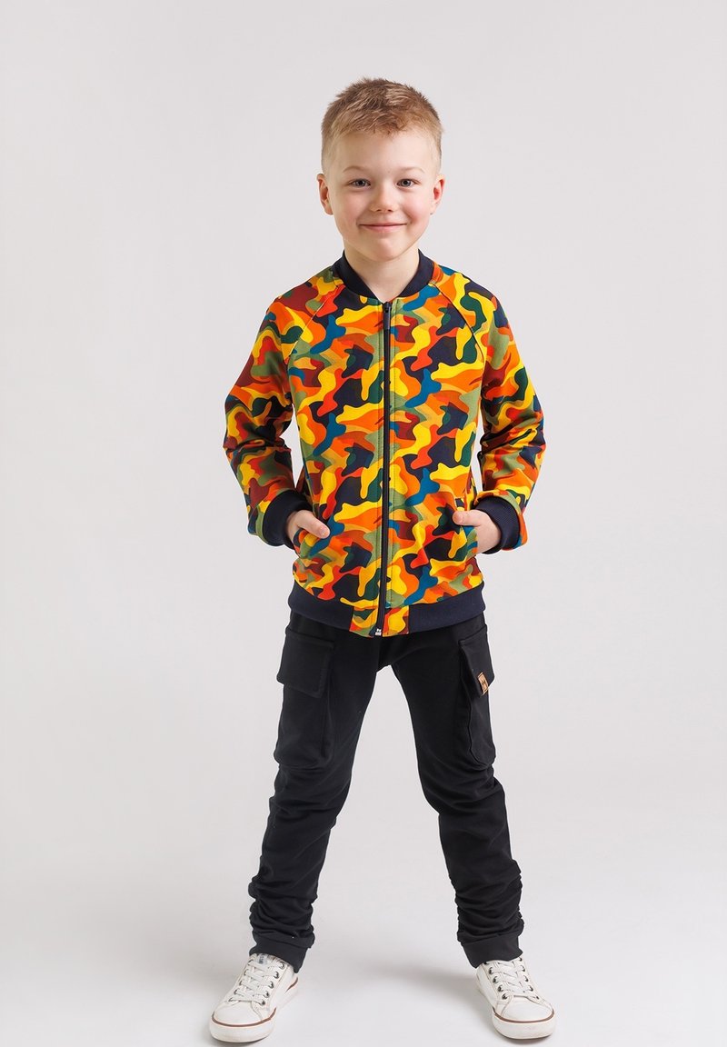 Chłopiec stoi z rękami w kieszeniach, ubrany w kolorową kurtkę w kamuflaż, czarne spodnie cargo i białe sneakersy na jednolitym tle.