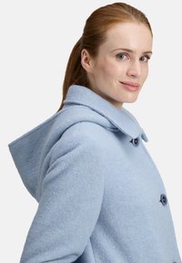 Donna con i capelli rossi raccolti, che indossa un cappotto di lana blu chiaro con cappuccio e bottoni scuri, mentre guarda oltre la spalla sorridendo.