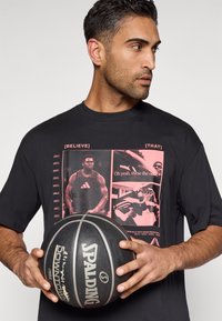 Must t-särk, millel on graafiline trükk korvpallimotiividega roosades toonides. Modell hoiab musta Spalding korvpalli. Lühikesed varrukad, ümar kaelus.