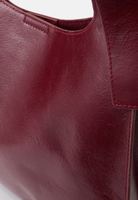 Geantă din piele bordo cu o textură netedă, având cusături vizibile și un design simplu, cu mânere curbate. Fără accesorii suplimentare.