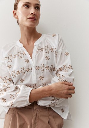 Vrouw draagt een witte blouse met bruine bloemige borduursels en beige broek, staand met gekruiste armen tegen een effen muur.