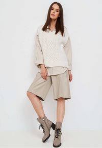 Maglione a trecce crema, camicia oversize beige, pantaloni corti marroni a pieghe e stivaletti taupe con lacci e suola nera. Modello in posa in piedi.
