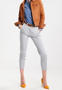Pantaloni grigi chiari su misura, camicia di denim e giacca in suede marrone. Tacchi alti gialli e cintura marrone aggiungono contrasto all'outfit.