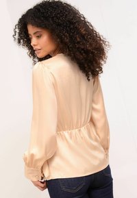 Svetlo broskyňová saténová blúzka s dlhými rukávmi, elastickým pásom a gombíkovými manžetami. Vyznačuje sa hladkou textúrou a jemným leskom.