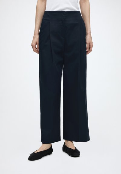 Femme portant un pantalon noir à jambes larges avec des ballerines noires, debout devant un fond blanc uni.