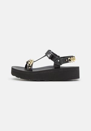 Sort platformsandal med tyk sål, justerbare remme og guldkæde detaljer på for- og bagremme, på ens hvid baggrund.