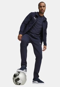 Marineblå atletisk træningstøj i blødt materiale, med en zip-up jakke, lange bukser og Puma-logo detaljer. Model står på en fodbold.