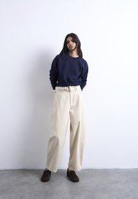 Γυναίκα που στέκεται μπροστά σε λευκό τοίχο, φοράει μπλε σκουρό πουλόβερ, μπεζ παντελόνια φαρδιάς γραμμής με ψηλή μέση και ζώνη, και σκούρα καφέ μοκασίνια.