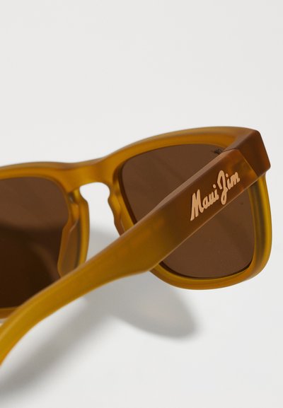 Brune nuancerede solbriller med en gennemsigtig amber-ramme. Funktionerne inkluderer en unik form og et indgraveret "Maui Jim"-logo på armen.