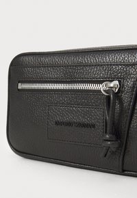 Fekete bőr pénztárca, cipzáros záródással, texturált felülettel, ezüst színű fémrészekkel és beleégett logóval. Téglalap alakú, két rekesszel.