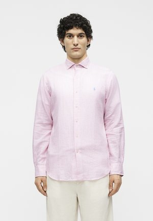 Άνδρας με σκούρα σγουρά μαλλιά, φοράει ένα ανοιχτό ροζ ριγέ πουκάμισο με μακριά μανίκια και ανοιχτόχρωμο παντελόνι, στέκεται μπροστά σε ένα μονόχρωμο φόντο.