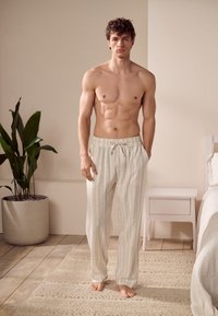 Pantalon de pyjama léger de couleur crème avec des rayures verticales, taille à cordon et texture douce, porté par un mannequin torse nu.