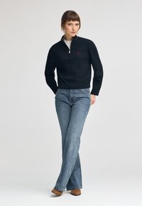 Svart stickad tröja med halvdragkedja och röd logotyp, kombinerad med ljusblå utsvängda jeans och bruna ankelskors.