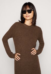 Brown ribbed knit dress with long sleeves and a crew neckline. Model wears black square sunglasses and displays silver rings on hands.