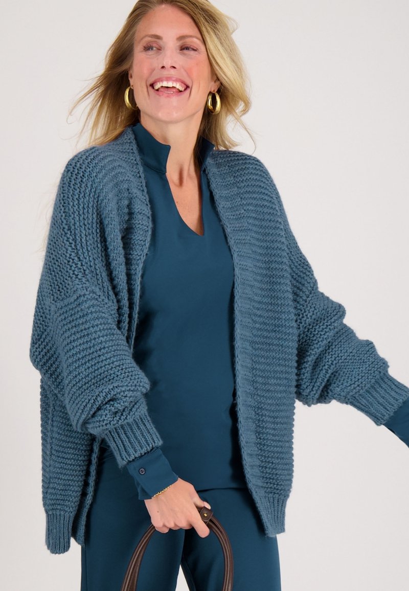 Chunky gebreide cardigan in blauw, met oversized mouwen en ribgebreide manchetten. Draagt over een teal top, vastgehouden met een bruine tas hengsel.