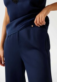 Femme ajustant un pantalon satiné bleu marine avec un haut sans manches assorti, montrant un gros plan des détails de la taille et des mains.