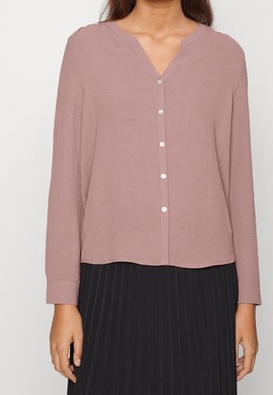 Woman wearing a long-sleeve mauve button-up blouse paired with a black pleated skirt, standing against a plain background.