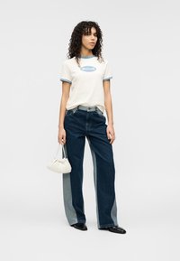 Λευκό μπλουζάκι με μπλε περίγραμμα και λογότυπο, συνδυασμένο με σκούρο τζιν παντελόνι με ανοιχτό μπλε πλαϊνά πάνελ, και μια μπεζ υφασμάτινη τσάντα.