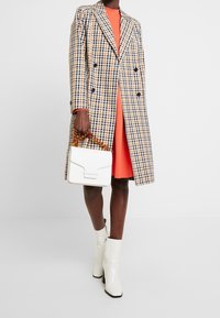 Kostymjacka med rutig mönster i blått och gult, orange klänning, vita lackläder ankle boots, och vit handväska med sköldpaddsformat handtag.