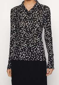Chemise noire à manches longues avec un motif abstrait blanc, col boutonné, fabriquée en tissu léger. Elle possède des boutons noirs et une coupe slim.