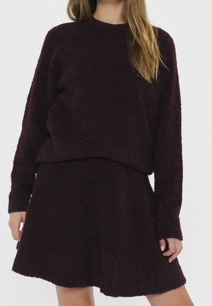 Femme portant un pull à manches longues violet foncé et duveteux ainsi qu'une jupe trapèze assortie, sur un fond blanc uni.