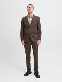 Brūns uzvalks ar piegriezumu, kas akcentē figūru, ar vienu pogu, ar ieliekamām atlokām un sānu kabatām. Valkāts virs gaiši bēša krekla un kombinēts ar melnām kurpēm.