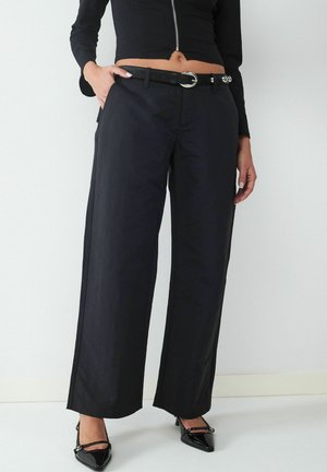 Zwarte wijde broek van gladde stof, met een ceintuur in de taille met een zilveren gesp en zijzakken, gestyled met zwarte puntige hakken.