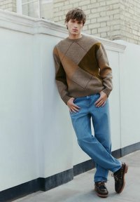 Brown and beige patterned sweater with diagonal design, light blue jeans, and dark brown shoes with laces and textured soles against a white wall.