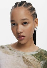 Model met gevlochten haar die een t-shirt met patroon draagt. Opvallende zilveren hartvormige oorbel met een parelaccent in het linkeroor.