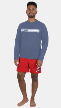 Blå sweatshirt med vit text "VIVID" och horisontella ränder, matchat med röda badshorts med palmträdsmönster. Barfota.