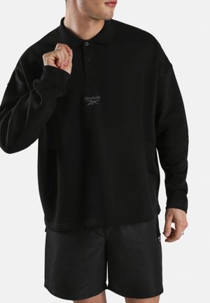Man draagt een zwarte poloshirt met lange mouwen met Reebok-logo en zwarte shorts, houdt de kraag van het shirt met één hand vast.
